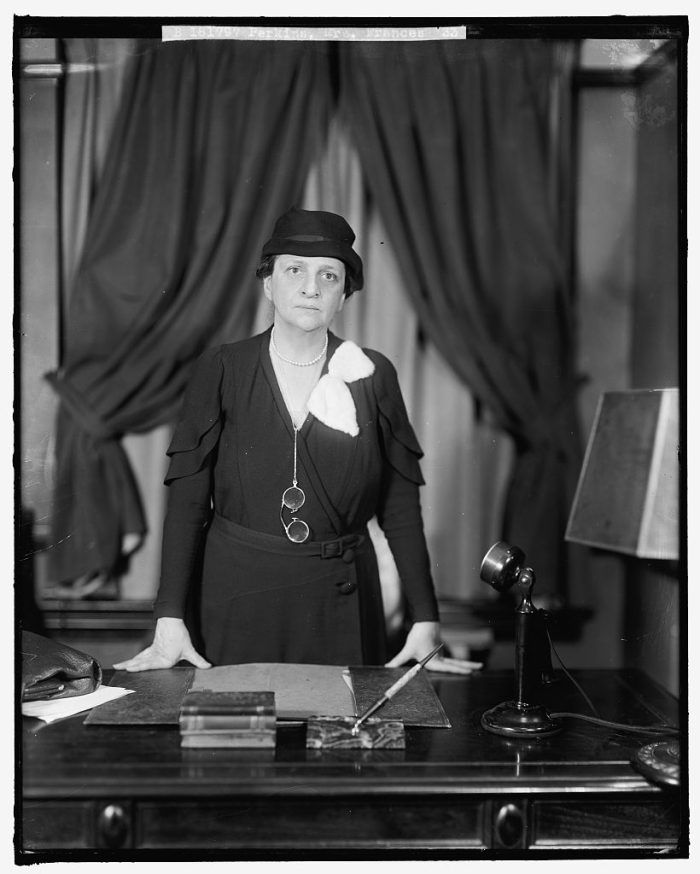 Black and white historical photograph of Frances Perkins standing behind a desk. She is dressed in a dark dress featuring a white collar, a belt at the waist, and a dark hat with a brim. She has round spectacles hanging from a chain around her neck and her expression is serious. On the desk, there are several books stacked to one side, a pen, and a classic black rotary dial telephone.
