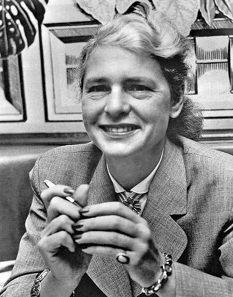 Black and white photograph of Margaret Bourke-White smiling, with crossed hands and wearing a suit with a leaf-patterned brooch.