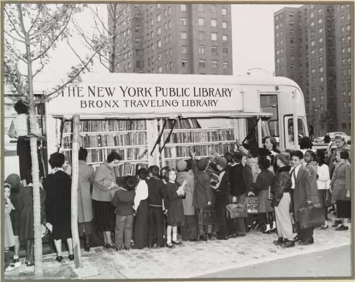 This black-and-white photograph shows a large group of children and adults gathered around a bookmobile, labeled "The New York Public Library - Bronx Traveling Library." The bookmobile's side is open, displaying rows of neatly arranged books. The scene is lively, with people browsing the books, while a librarian assists. The setting is in an urban area, with tall apartment buildings visible in the background. Some children carry school bags, and a few are dressed in coats, suggesting it may be a chilly day.