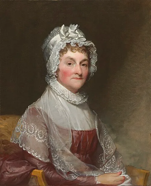 Oil portrait of Abigail Adams painted around 1810. She sits facing slightly left, wearing a lace cap tied with a white ribbon, a sheer shawl over a red dress, and a calm, dignified expression. The background is dark and plain, highlighting her face and attire.