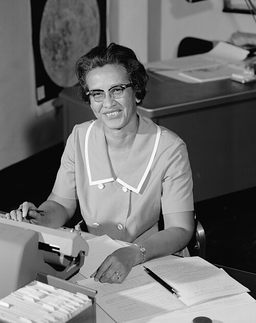 Katherine Johnson sits at her desk at NASA’s Langley Research Center, smiling while working with papers and a typewriter. She wears glasses and a light-colored dress with a white collar; behind her is a desk with documents and a large map of the Moon on the wall.