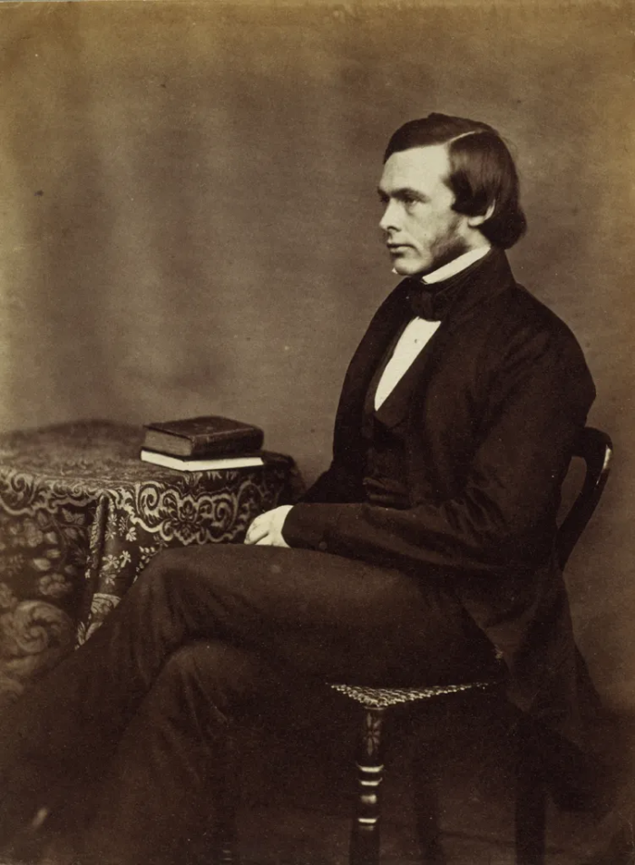Sepia-toned photograph of a young Dr. Joseph Lister seated in profile. He is a clean-shaven man with dark hair and prominent sideburns, wearing a dark frock coat, waistcoat, and bow tie. He sits with hands clasped in his lap beside a small table draped in patterned cloth, on which two cloth-bound books rest.