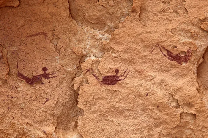 Three small human figures painted in reddish-brown pigment on a sandstone cave wall, limbs bent and splayed as if swimming.